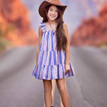 A young girl wearing the Mabel and Honey Riley Rose Dress smiles on a road, paired with a brown cowboy hat and boots. Red rock formations blur in the background.