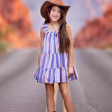 A young girl wearing the Mabel and Honey Riley Rose Dress smiles on a road, paired with a brown cowboy hat and boots. Red rock formations blur in the background.
