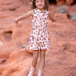 A young child with curly hair smiles and spreads their arms on red rock terrain, wearing the sleeveless collared Mavery Floral Romper by Mabel and Honey with light-colored boots.