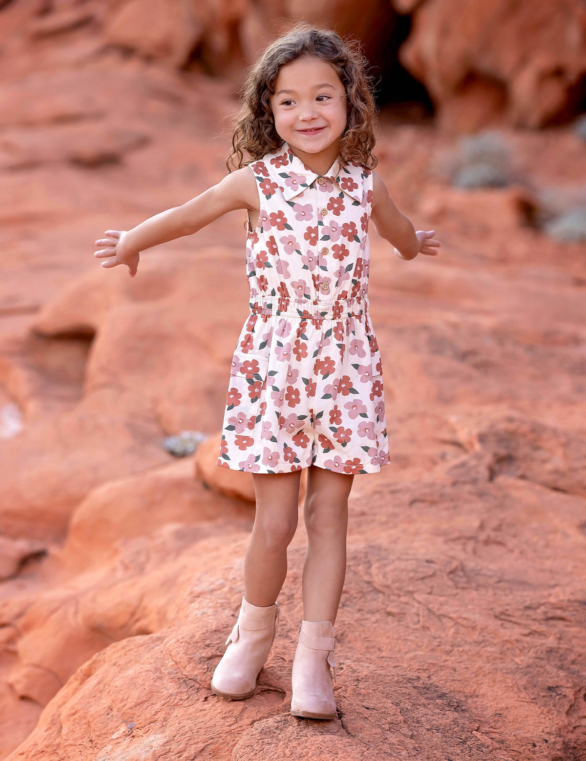 A young child with curly hair smiles and spreads their arms on red rock terrain, wearing the sleeveless collared Mavery Floral Romper by Mabel and Honey with light-colored boots.