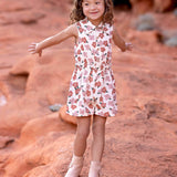 A young child with curly hair smiles and spreads their arms on red rock terrain, wearing the sleeveless collared Mavery Floral Romper by Mabel and Honey with light-colored boots.