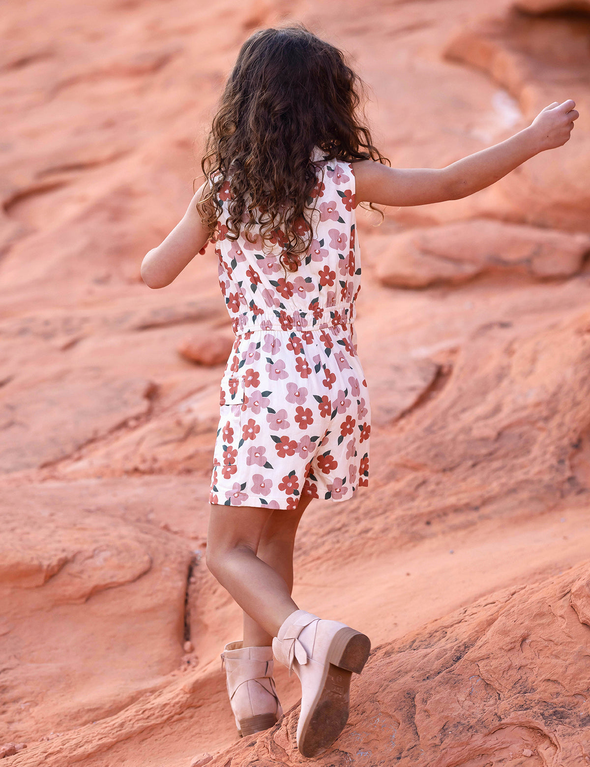 A young girl with curly hair, seen from behind, walks on reddish rocks wearing the Mavery Floral Romper by Mabel and Honey with an elastic waist, paired with light pink boots.