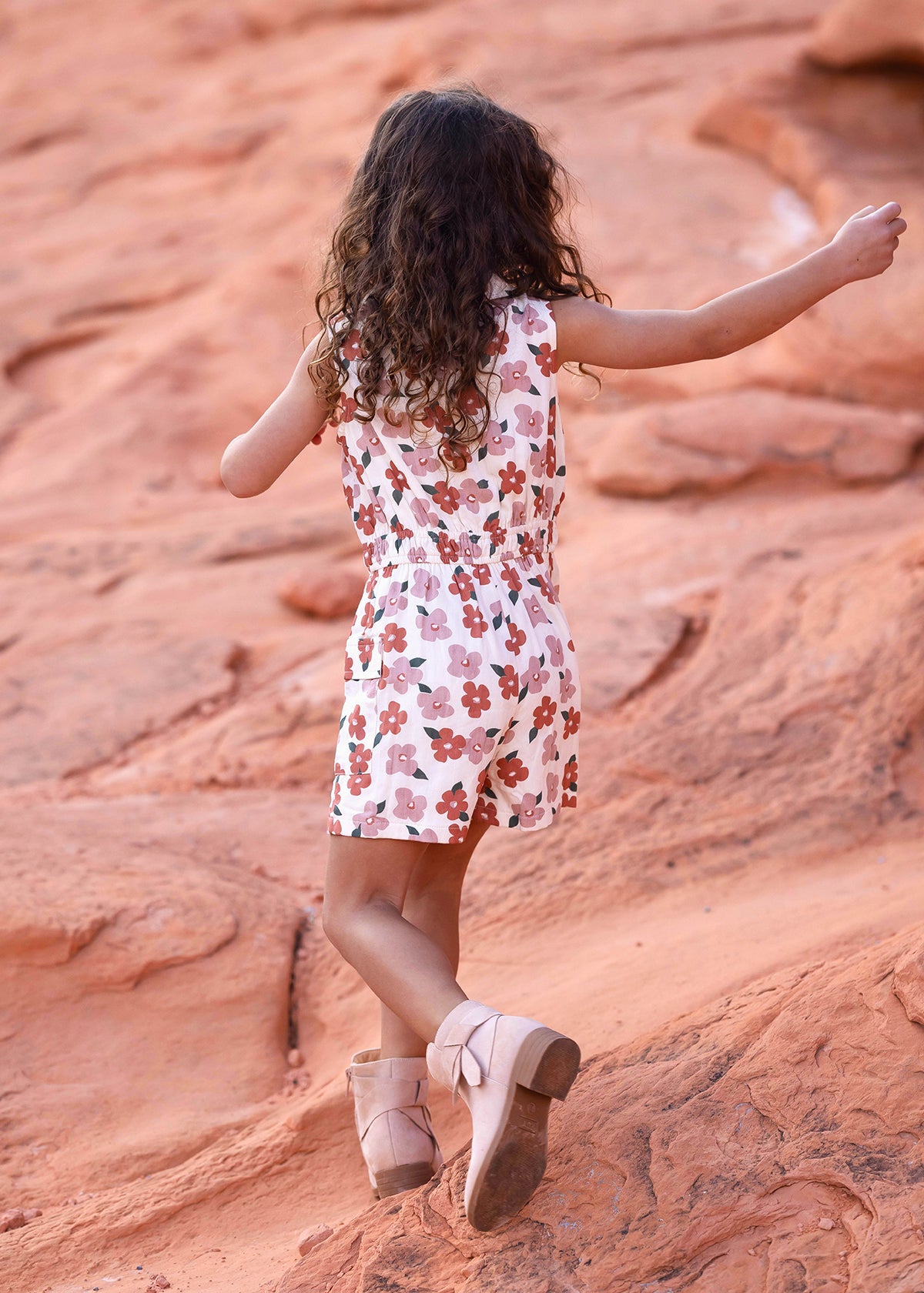 A young girl with curly hair, seen from behind, walks on reddish rocks wearing the Mavery Floral Romper by Mabel and Honey with an elastic waist, paired with light pink boots.