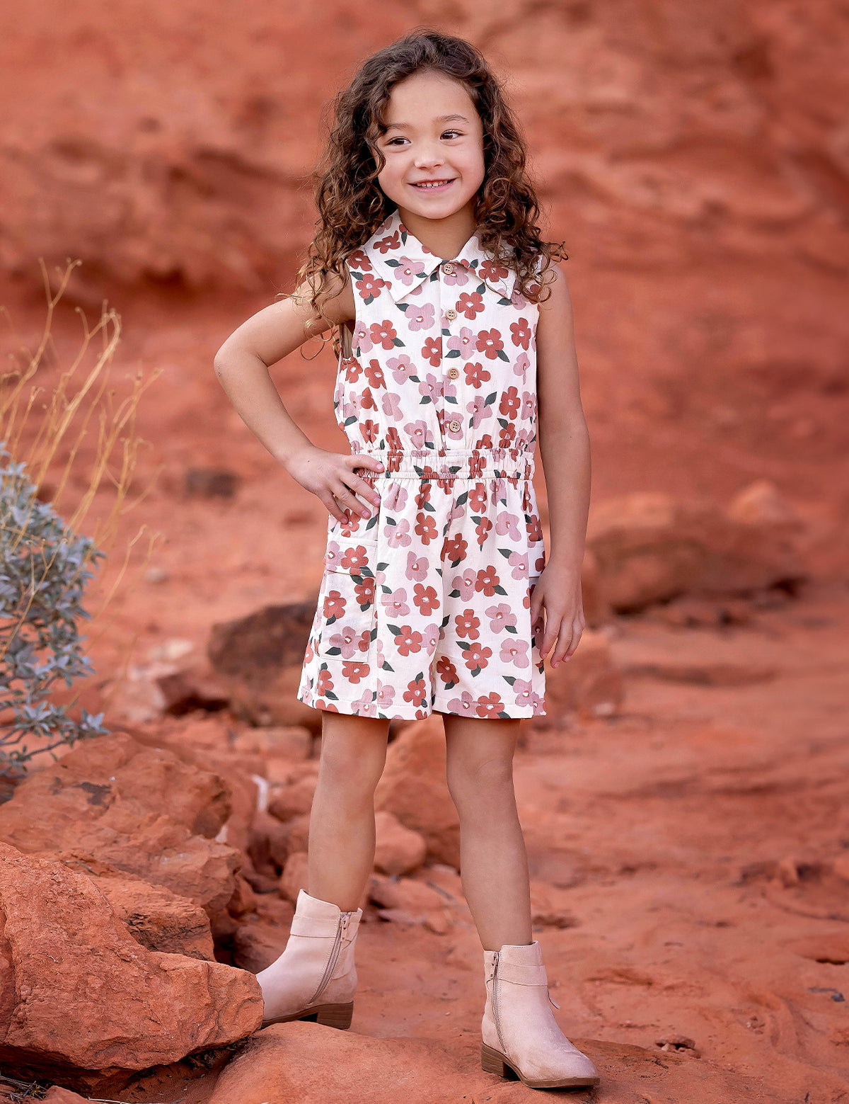 A young girl with curly hair smiles on red rocks, wearing the Mavery Floral Romper by Mabel and Honey with light tan ankle boots, set against a desert landscape of reddish soil and sparse vegetation.