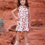 A young girl with curly hair smiles on red rocks, wearing the Mavery Floral Romper by Mabel and Honey with light tan ankle boots, set against a desert landscape of reddish soil and sparse vegetation.