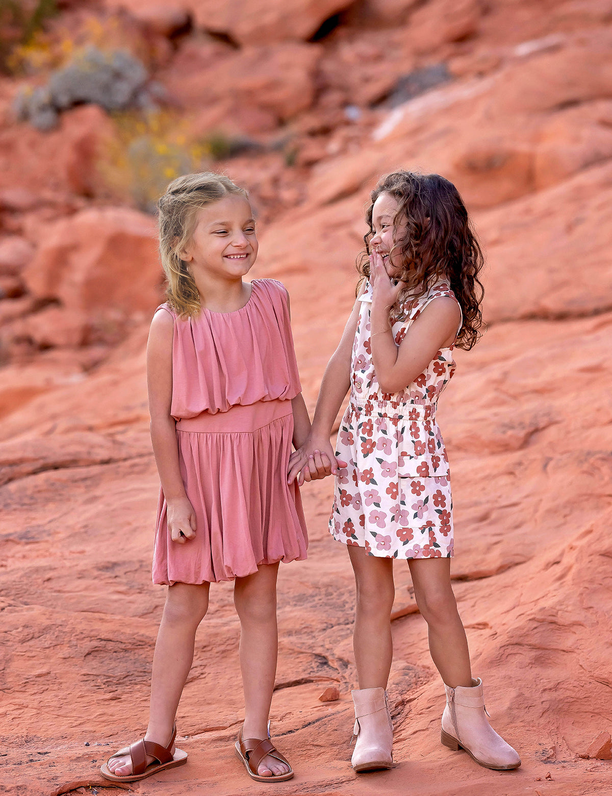 Two young girls stand on reddish rocks, smiling and holding hands. One wears a pink dress and brown sandals; the other is in a Mavery Floral Romper by Mabel and Honey with beige boots. They look happy and playful outdoors.