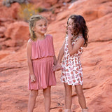 Two young girls stand on reddish rocks, smiling and holding hands. One wears a pink dress and brown sandals; the other is in a Mavery Floral Romper by Mabel and Honey with beige boots. They look happy and playful outdoors.