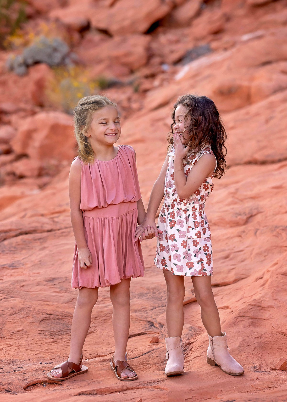 Two young girls stand on reddish rocks, smiling and holding hands. One wears a pink dress and brown sandals; the other is in a Mavery Floral Romper by Mabel and Honey with beige boots. They look happy and playful outdoors.