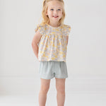 A young blonde girl smiles in the Mila Two Piece Set by Mabel and Honey, featuring a pastel-print top and sage green shorts, paired with sparkly shoes against a plain white background.