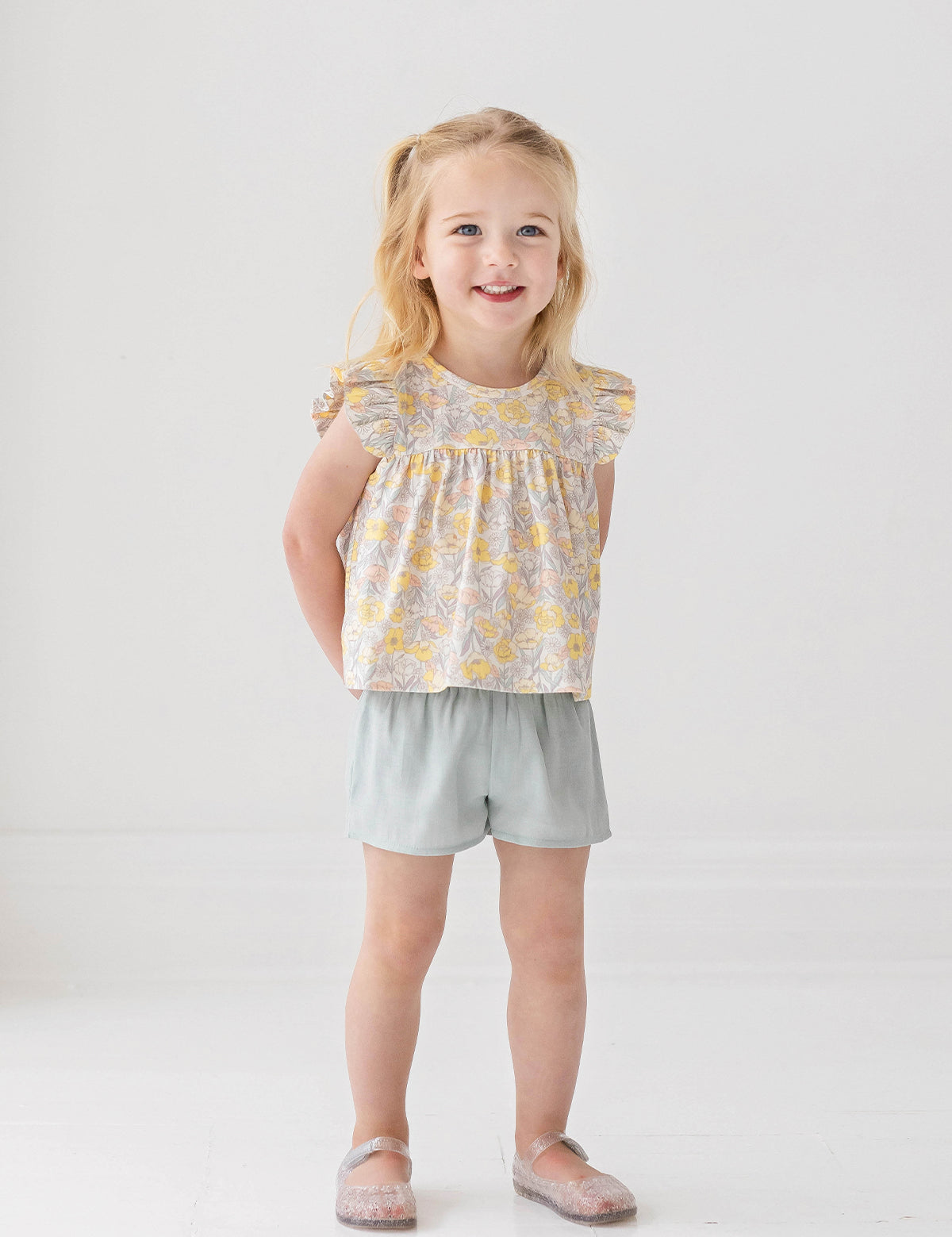 A young blonde girl smiles in the Mila Two Piece Set by Mabel and Honey, featuring a pastel-print top and sage green shorts, paired with sparkly shoes against a plain white background.