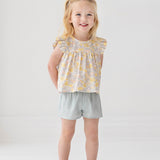 A young blonde girl smiles in the Mila Two Piece Set by Mabel and Honey, featuring a pastel-print top and sage green shorts, paired with sparkly shoes against a plain white background.
