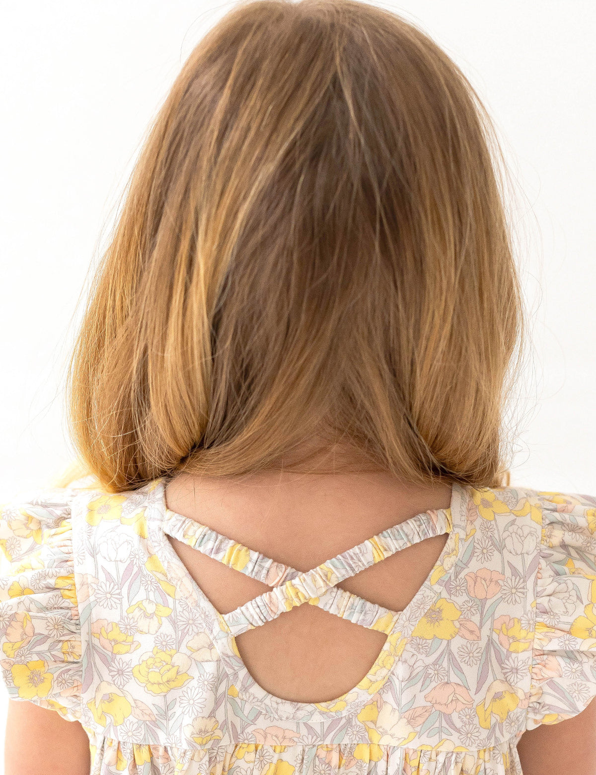 A young girl with light brown hair, seen from behind, wears the Mila Two Piece Set by Mabel and Honey—sage green shorts and a floral-patterned top with yellow and pink flowers plus crossed back straps.