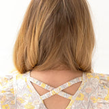 A young girl with light brown hair, seen from behind, wears the Mila Two Piece Set by Mabel and Honey—sage green shorts and a floral-patterned top with yellow and pink flowers plus crossed back straps.