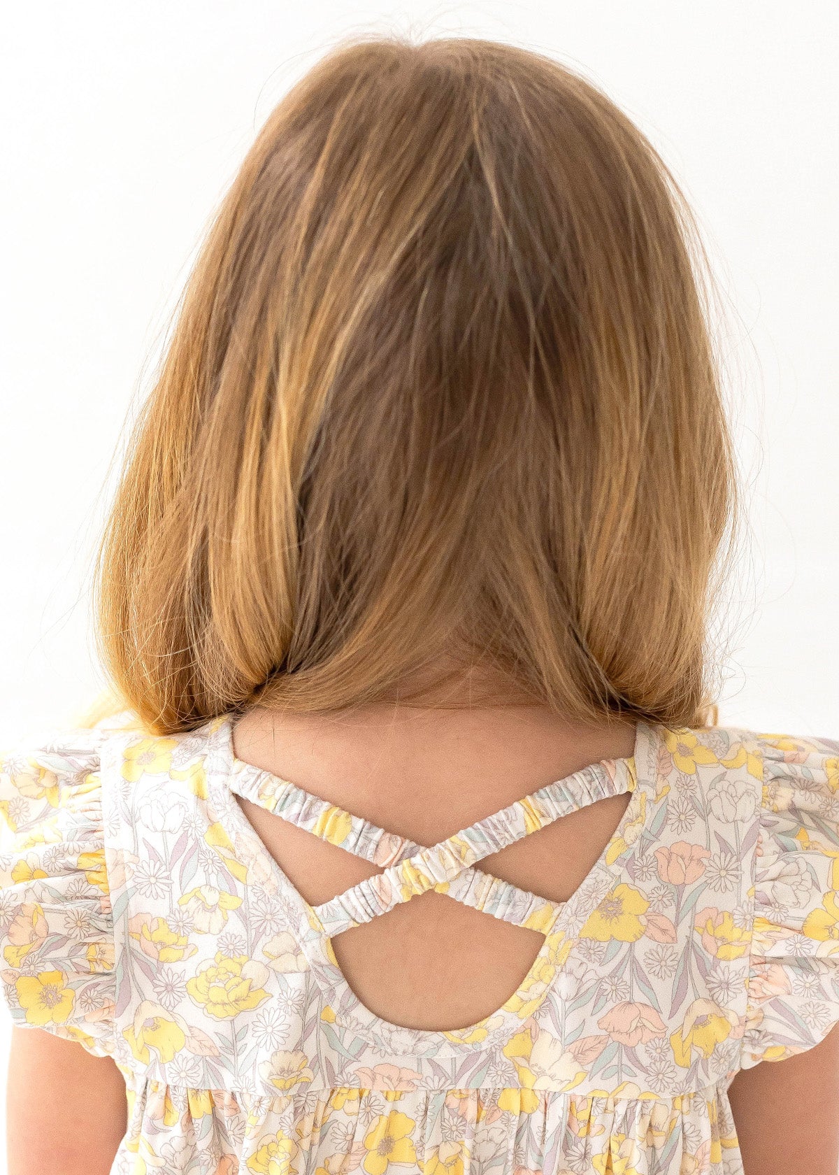 A young girl with light brown hair, seen from behind, wears the Mila Two Piece Set by Mabel and Honey—sage green shorts and a floral-patterned top with yellow and pink flowers plus crossed back straps.