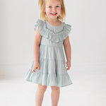 A young child with blonde pigtails smiles while standing against a white background, wearing the Mabel and Honey Mila Green Emerald Dress.