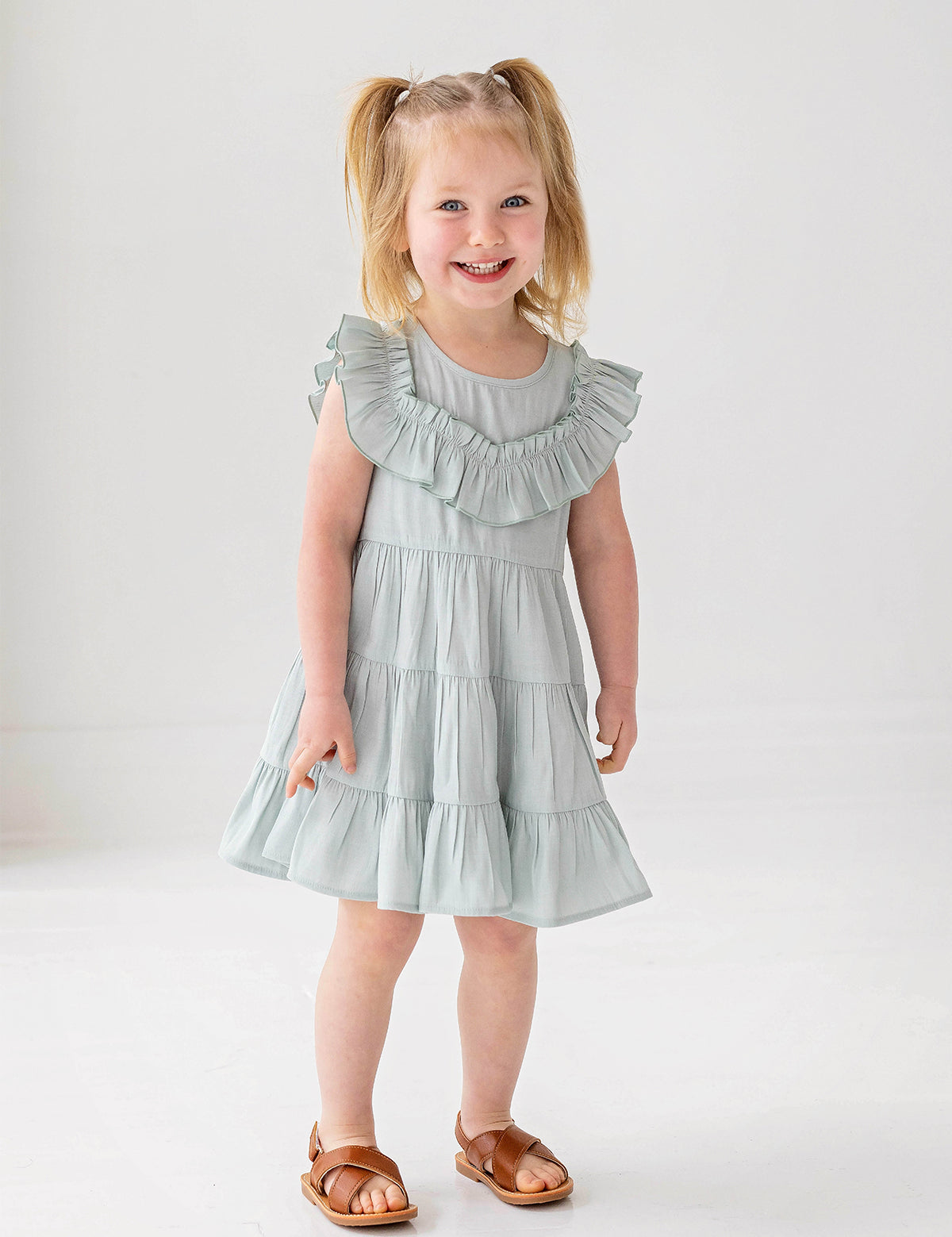 A young child with blonde pigtails smiles while standing against a white background, wearing the Mabel and Honey Mila Green Emerald Dress.