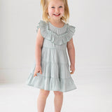 A young child with blonde pigtails smiles while standing against a white background, wearing the Mabel and Honey Mila Green Emerald Dress.