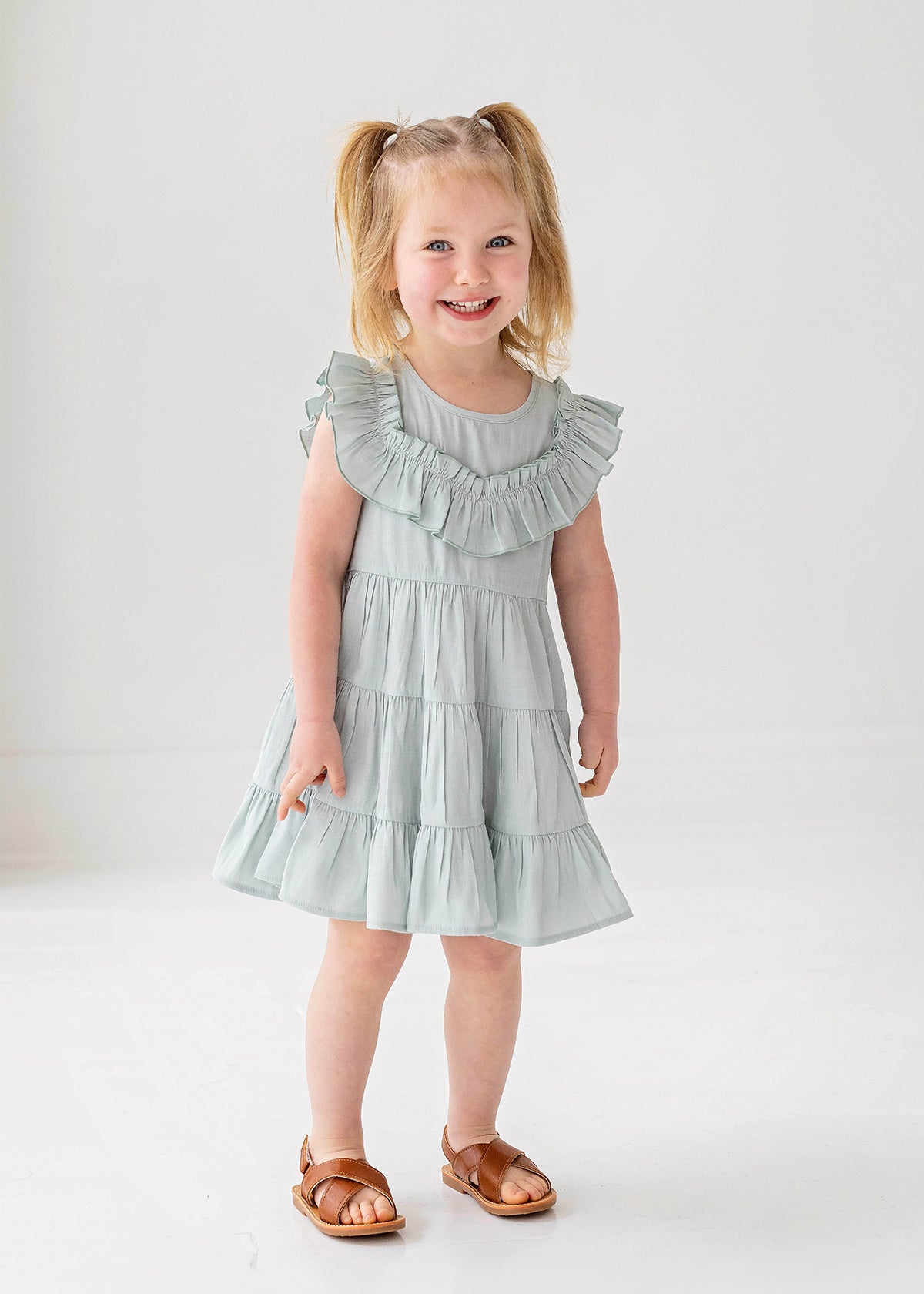 A young child with blonde pigtails smiles while standing against a white background, wearing the Mabel and Honey Mila Green Emerald Dress.