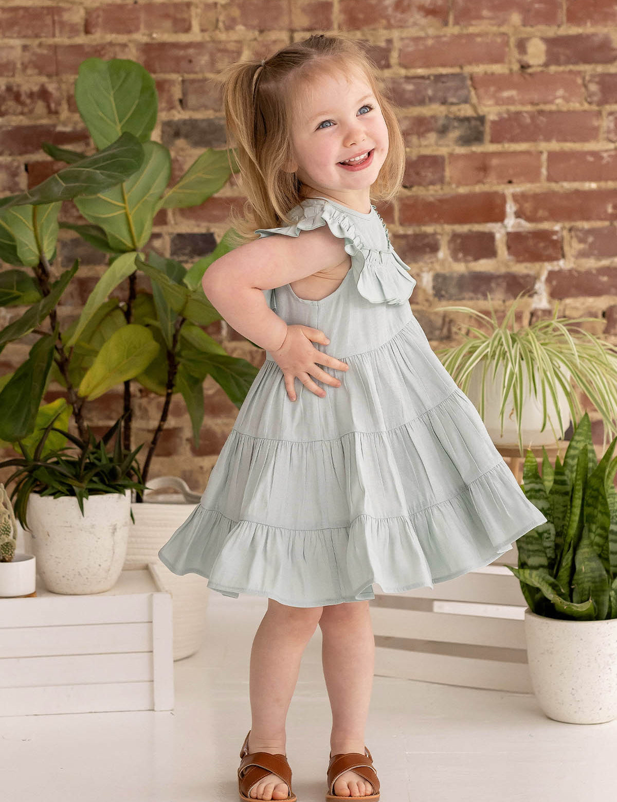 A young girl with blonde pigtails smiles while posing indoors in the Mila Green Emerald Dress by Mabel and Honey, paired with brown sandals near green potted plants and a brick wall.