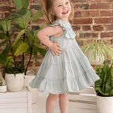 A young girl with blonde pigtails smiles while posing indoors in the Mila Green Emerald Dress by Mabel and Honey, paired with brown sandals near green potted plants and a brick wall.