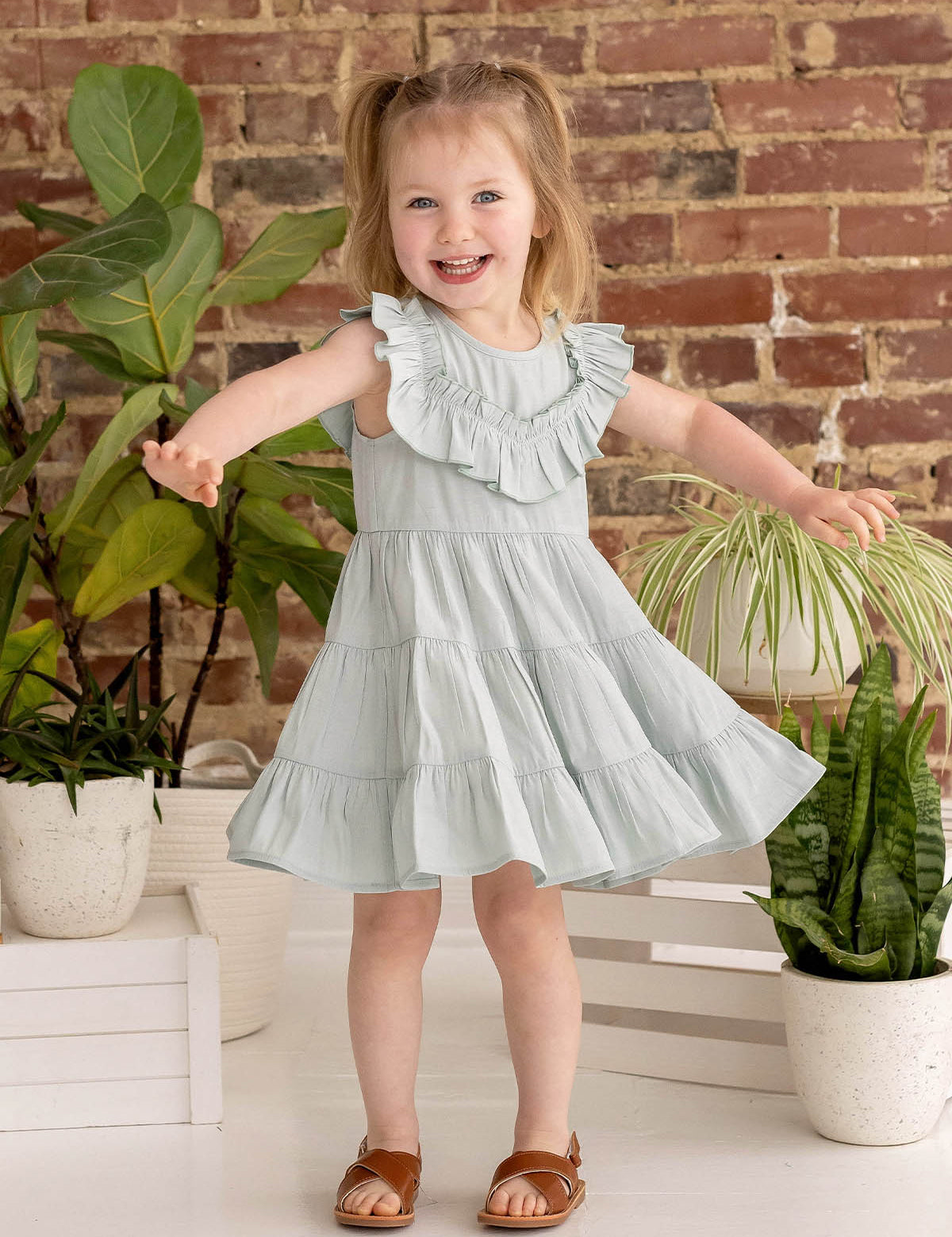 A smiling young girl with light brown pigtails wears the Mila Green Emerald Dress by Mabel and Honey, standing with arms outstretched among potted green plants against a rustic brick wall background, paired with brown sandals.