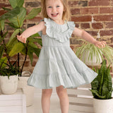 A smiling young girl with light brown pigtails wears the Mila Green Emerald Dress by Mabel and Honey, standing with arms outstretched among potted green plants against a rustic brick wall background, paired with brown sandals.