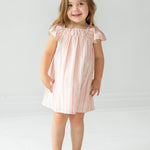 A young girl with long brown hair wears the Sandy Dress by Mabel and Honey, a light pink and white striped dress with flutter sleeves, paired with beige sandals, standing and smiling against a plain white background.
