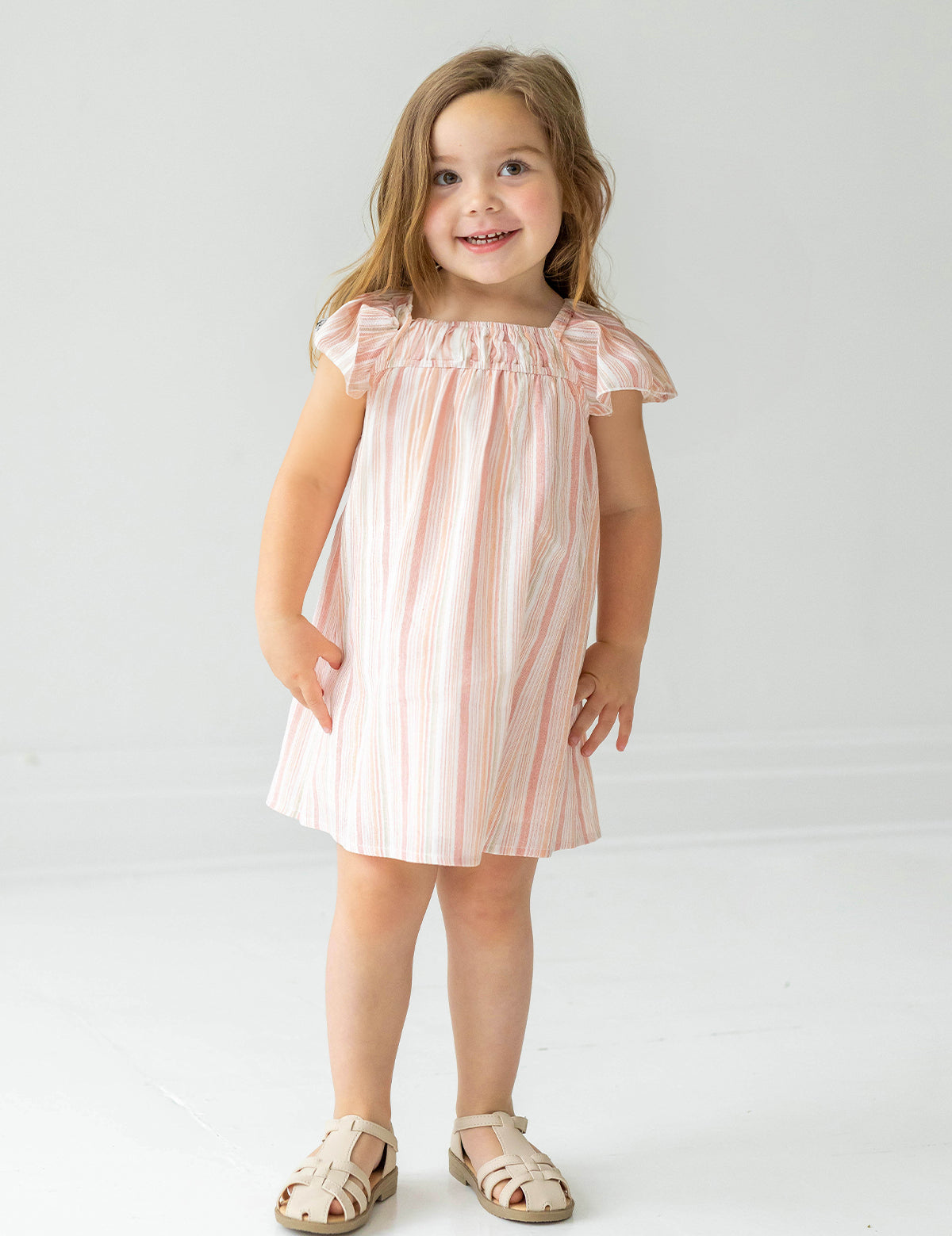A young girl with long brown hair wears the Sandy Dress by Mabel and Honey, a light pink and white striped dress with flutter sleeves, paired with beige sandals, standing and smiling against a plain white background.