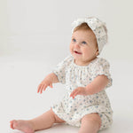 A smiling baby wears the Bluebell Baby Romper from Mabel and Honey with puff sleeves and a matching headband, sitting on the floor against a plain white background.