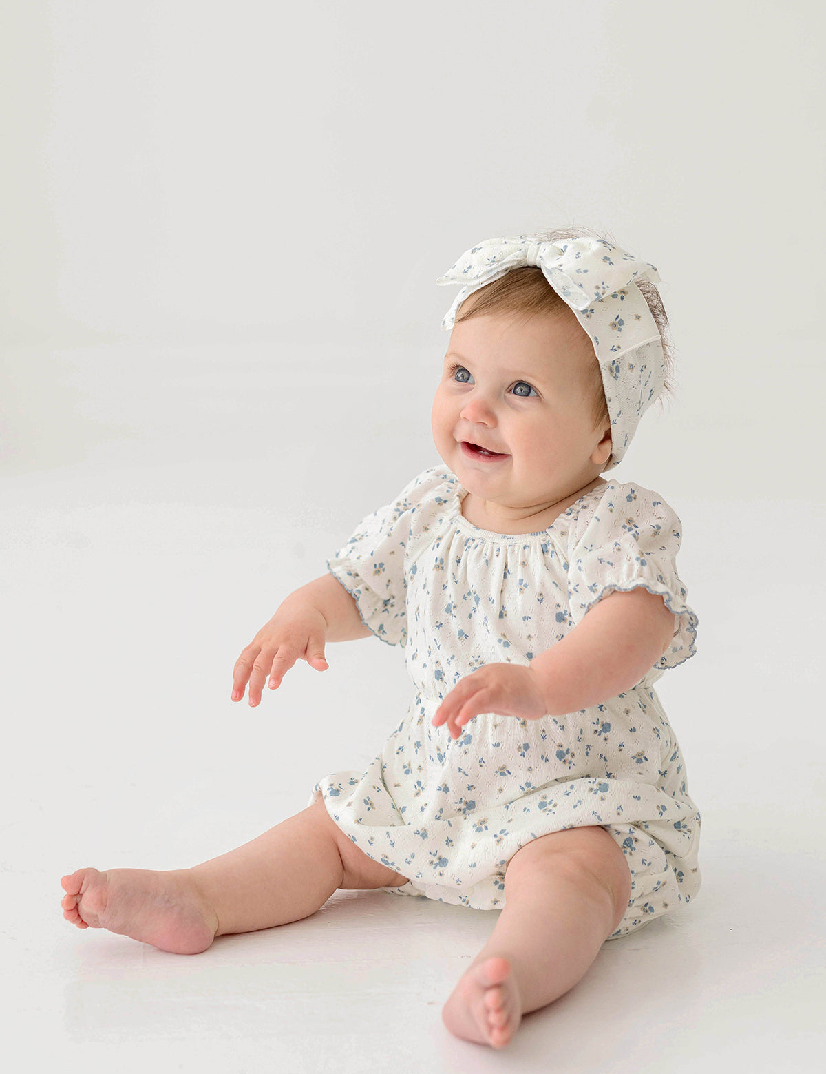 A smiling baby wears the Bluebell Baby Romper from Mabel and Honey with puff sleeves and a matching headband, sitting on the floor against a plain white background.
