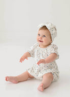 A smiling baby wears the Bluebell Baby Romper from Mabel and Honey with puff sleeves and a matching headband, sitting on the floor against a plain white background.