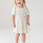 A young girl with light brown hair in pigtails smiles against a white background, wearing the Mabel and Honey Bluebells Drop Shoulder Dress with a blue floral print, along with white socks and shoes.