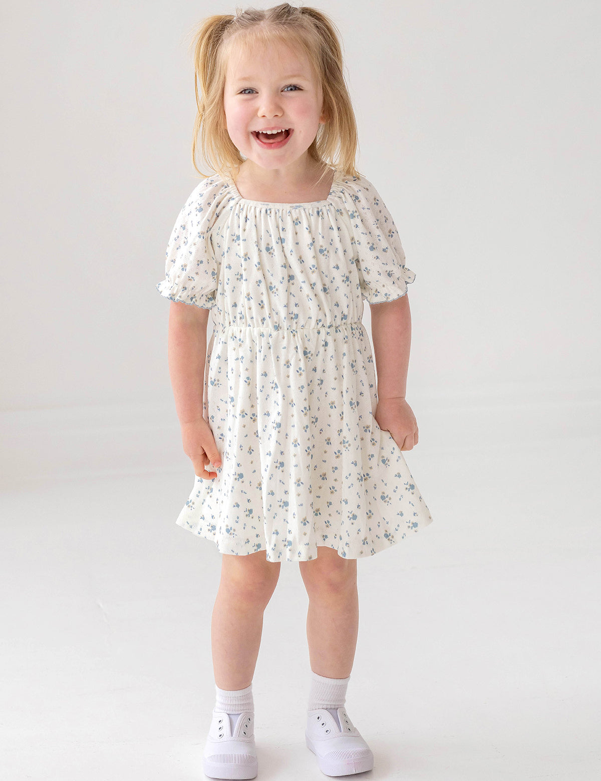 A young girl with light brown hair in pigtails smiles against a white background, wearing the Mabel and Honey Bluebells Drop Shoulder Dress with a blue floral print, along with white socks and shoes.