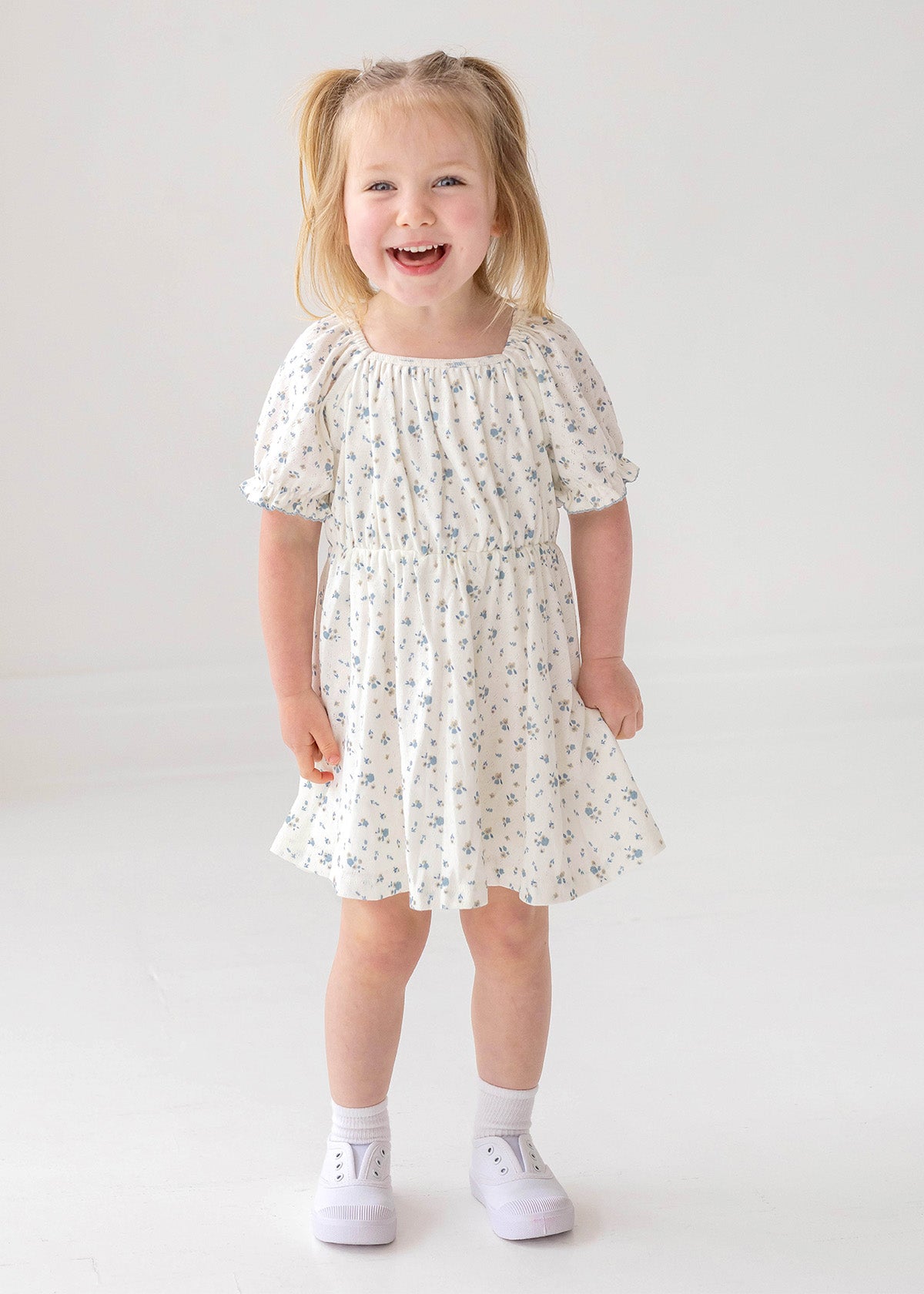 A young girl with light brown hair in pigtails smiles against a white background, wearing the Mabel and Honey Bluebells Drop Shoulder Dress with a blue floral print, along with white socks and shoes.