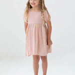 A young girl with long curly blonde hair smiles in the Mabel and Honey Cranberry Crush Dress—a peach and white striped, short-sleeve girls dress—paired with transparent sandals against a plain white background.