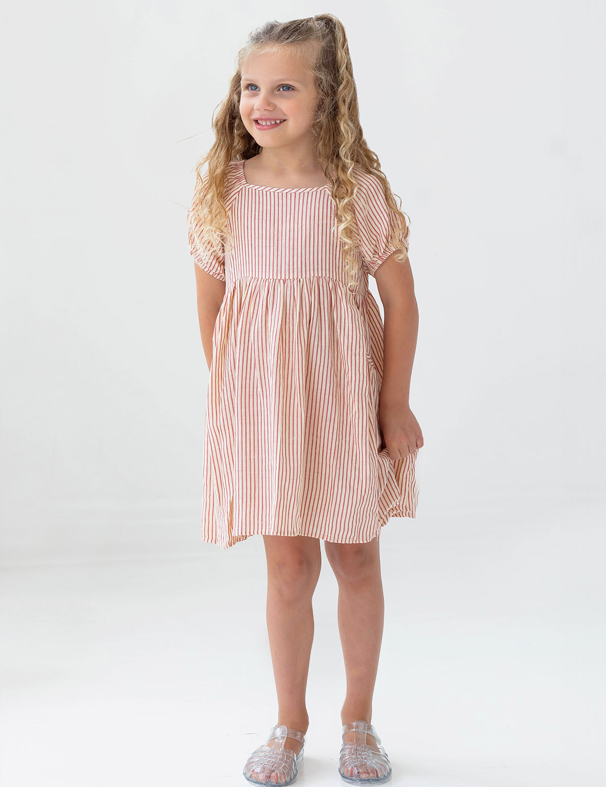 A young girl with long curly blonde hair smiles in the Mabel and Honey Cranberry Crush Dress—a peach and white striped, short-sleeve girls dress—paired with transparent sandals against a plain white background.