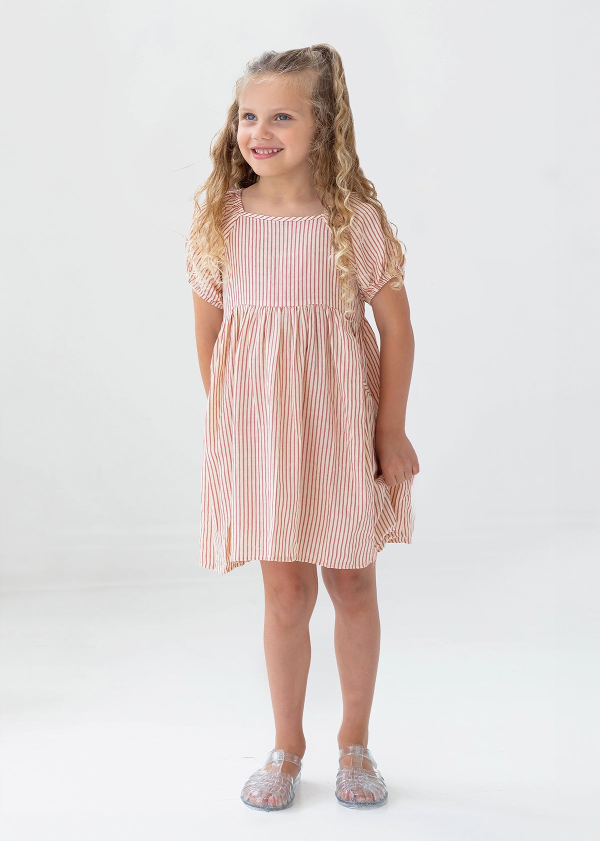 A young girl with long curly blonde hair smiles in the Mabel and Honey Cranberry Crush Dress—a peach and white striped, short-sleeve girls dress—paired with transparent sandals against a plain white background.