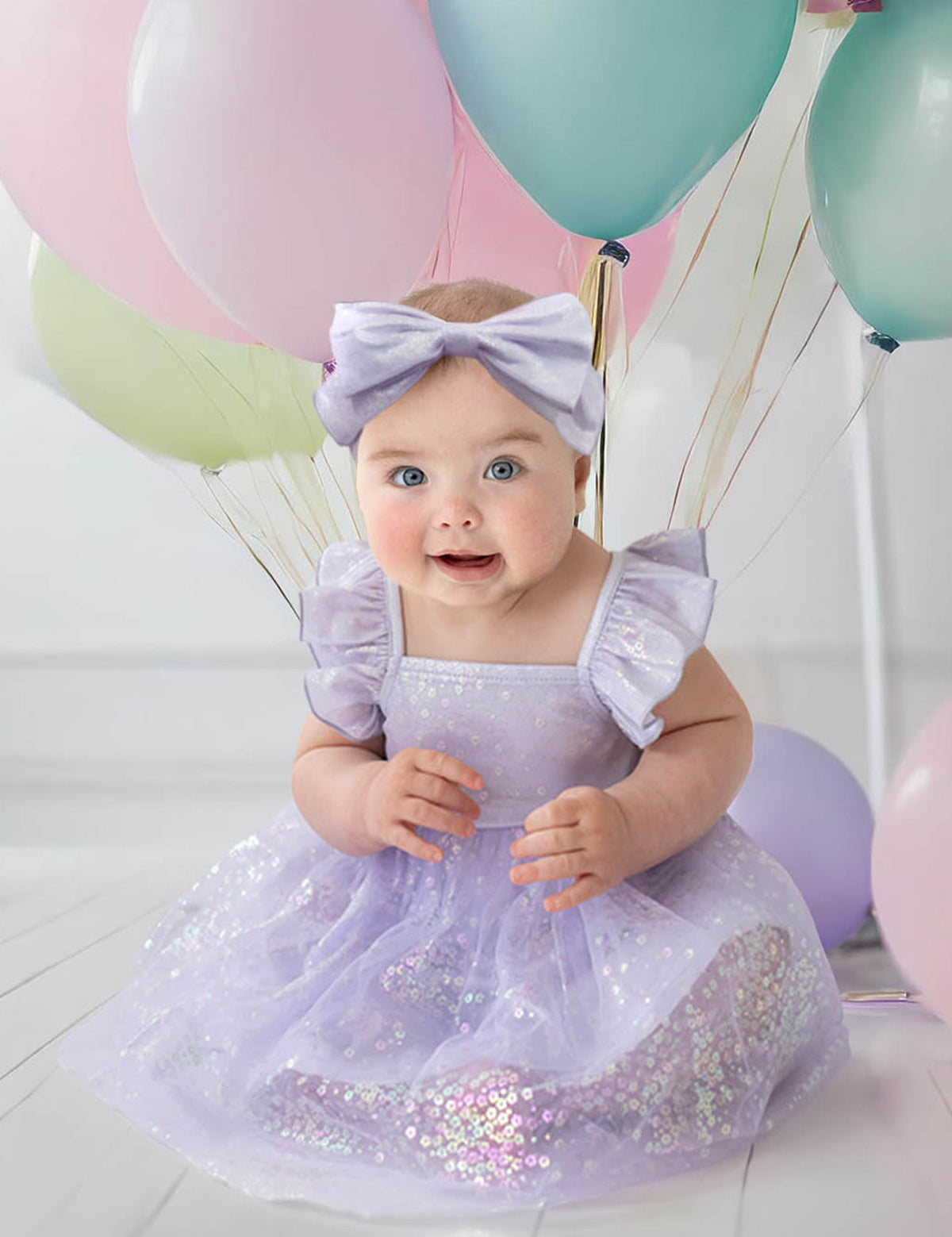 A baby in the Purple Star Empire Waist Dress by Isobella and Chloe, with a matching headband, sits on a white floor among pastel balloons. She smiles brightly, her cheerful charm enhanced by soft lighting in the background.