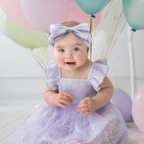 A baby in the Purple Star Empire Waist Dress by Isobella and Chloe, with a matching headband, sits on a white floor among pastel balloons. She smiles brightly, her cheerful charm enhanced by soft lighting in the background.