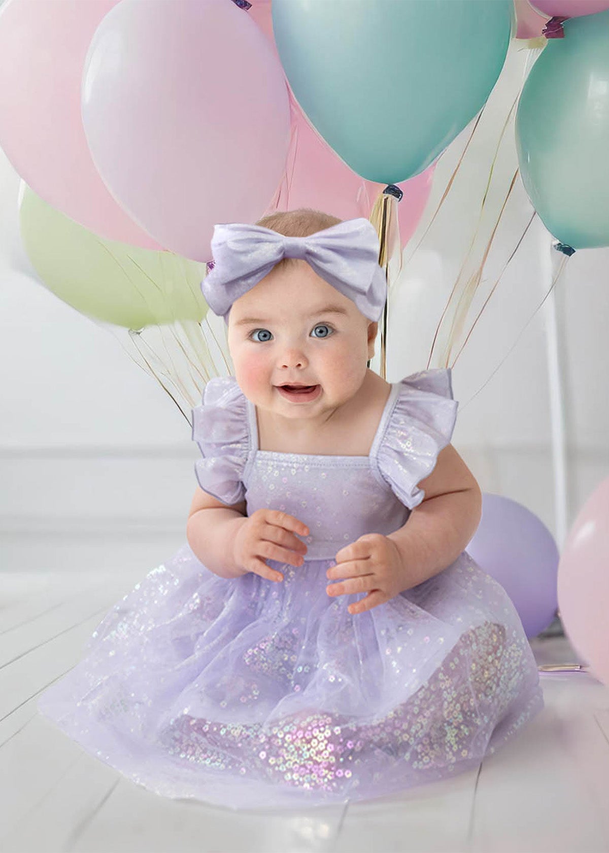 A baby in the Purple Star Empire Waist Dress by Isobella and Chloe, with a matching headband, sits on a white floor among pastel balloons. She smiles brightly, her cheerful charm enhanced by soft lighting in the background.