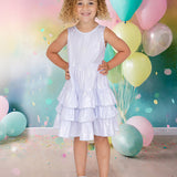 A young girl with curly blonde hair smiles, posing with hands on hips in the Isobella and Chloe Purple Star Glimmering Tiered Dress and sandals. She stands among pastel balloons and confetti, set against a colorful, festive background.