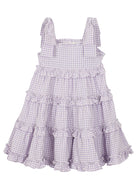 The Super Bloom Tiered Dress by Mabel and Honey is a sleeveless, knee-length lavender gingham dress with white checks, ruffled tiers, and wide shoulder straps tied in bows.