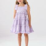 A young girl with long curly hair smiles in the Mabel and Honey Super Bloom Tiered Dress, featuring lavender gingham, ruffles, and bows. She pairs it with clear sandals while standing against a plain white background.
