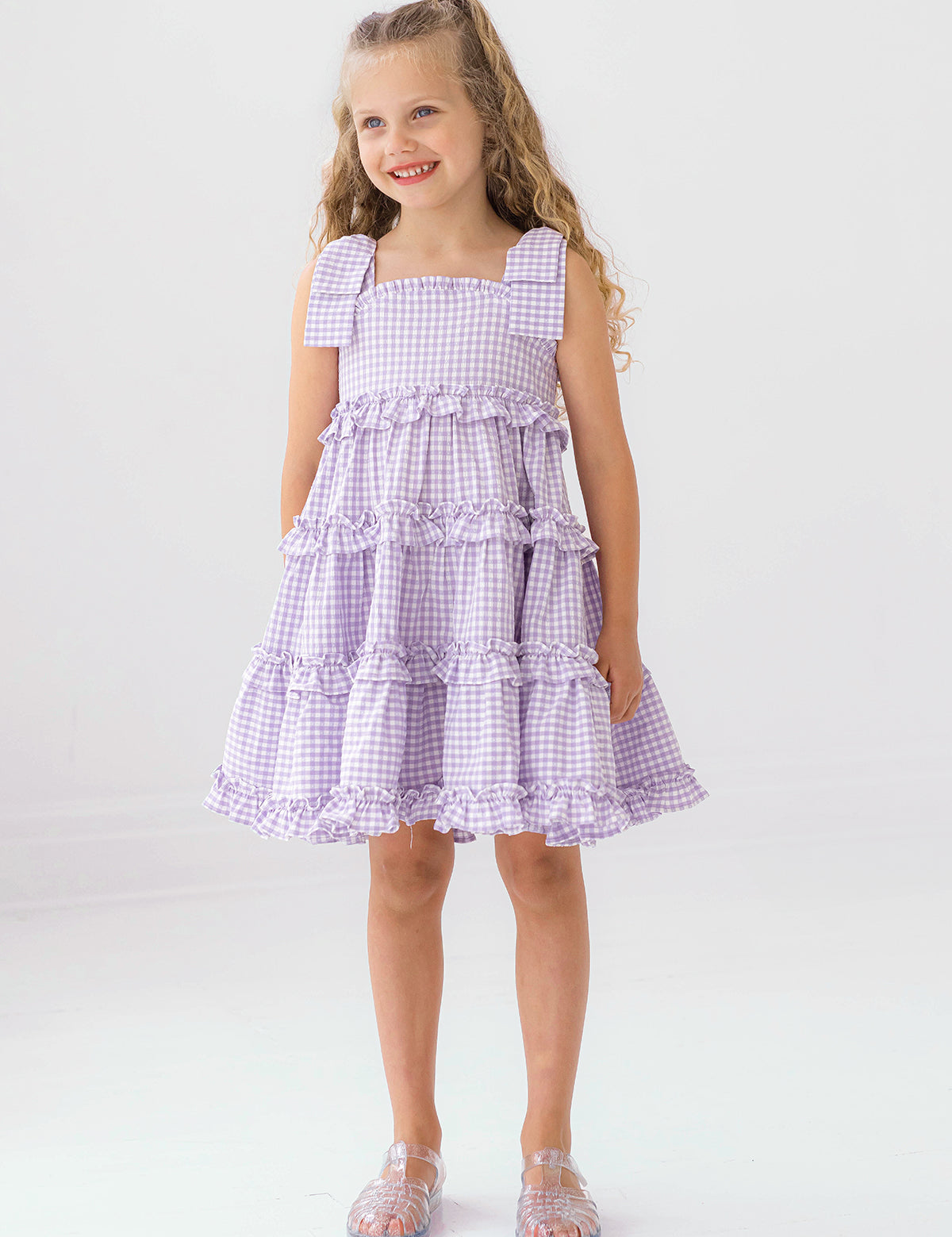 A young girl with long curly hair smiles in the Mabel and Honey Super Bloom Tiered Dress, featuring lavender gingham, ruffles, and bows. She pairs it with clear sandals while standing against a plain white background.