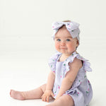 A blue-eyed baby sits on the floor in the Mabel and Honey Tiny Tulip Baby Romper, a lavender gauze outfit with ruffled sleeves and matching large bow headband, both featuring small floral patterns and pink tulips, against a white background.