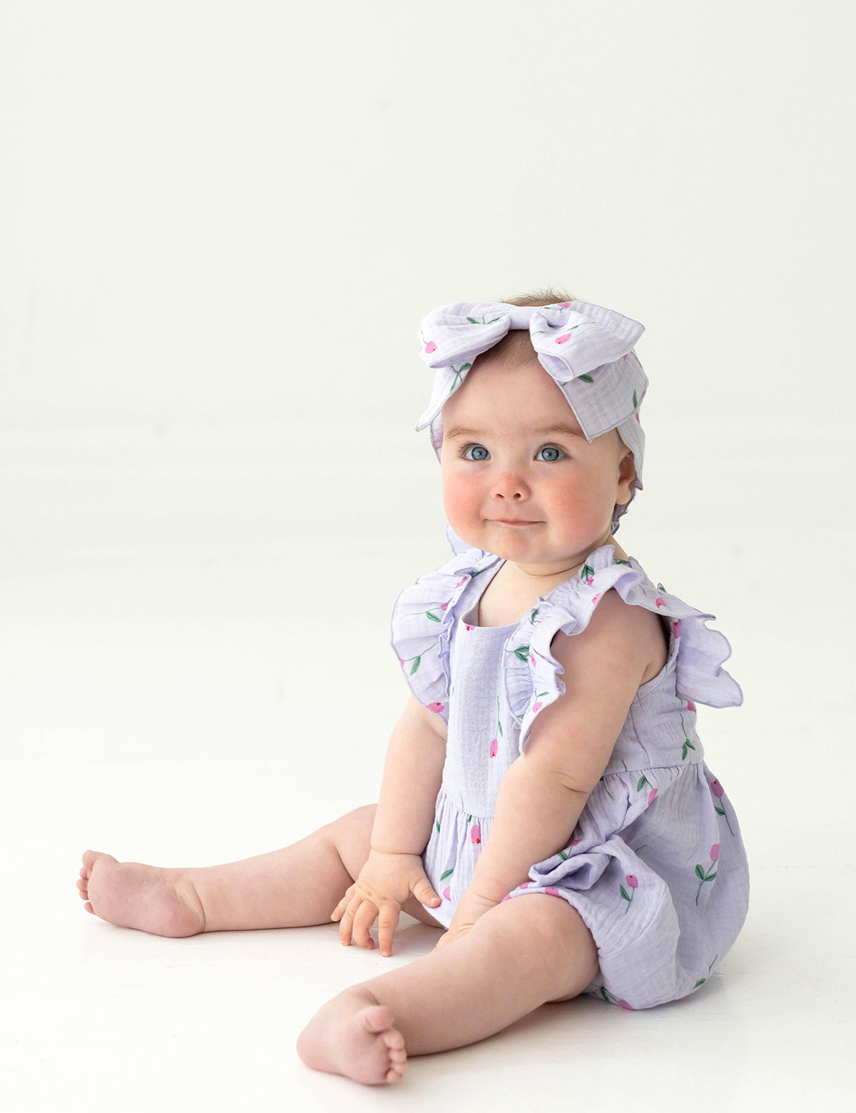 A blue-eyed baby sits on the floor in the Mabel and Honey Tiny Tulip Baby Romper, a lavender gauze outfit with ruffled sleeves and matching large bow headband, both featuring small floral patterns and pink tulips, against a white background.