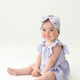 A blue-eyed baby sits on the floor in the Mabel and Honey Tiny Tulip Baby Romper, a lavender gauze outfit with ruffled sleeves and matching large bow headband, both featuring small floral patterns and pink tulips, against a white background.
