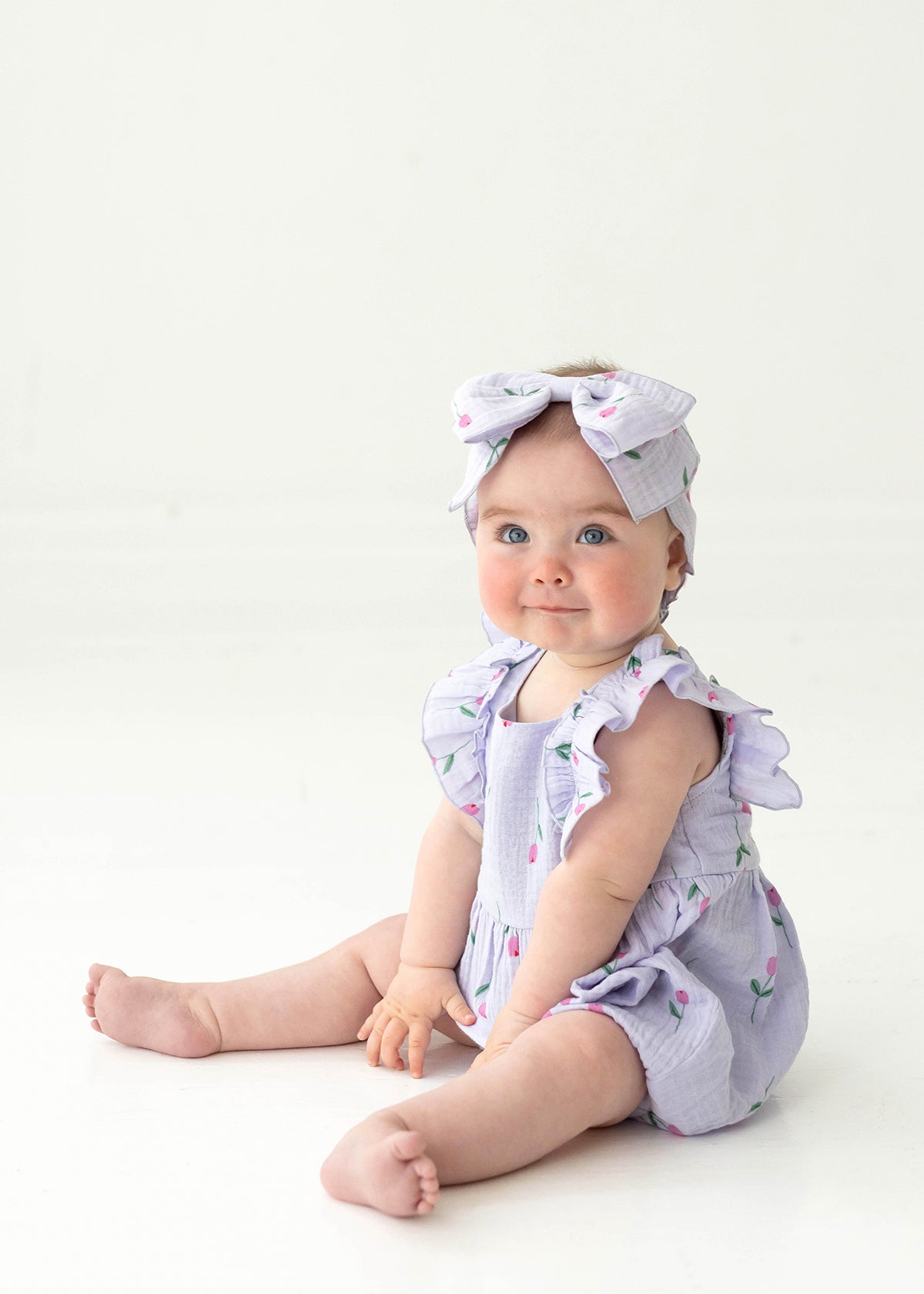 A blue-eyed baby sits on the floor in the Mabel and Honey Tiny Tulip Baby Romper, a lavender gauze outfit with ruffled sleeves and matching large bow headband, both featuring small floral patterns and pink tulips, against a white background.