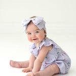 A smiling blue-eyed baby sits on the floor amid pink tulips, wearing the Mabel and Honey Tiny Tulip Baby Romper with ruffles and a matching cherry-print bow headband, set against a white background.