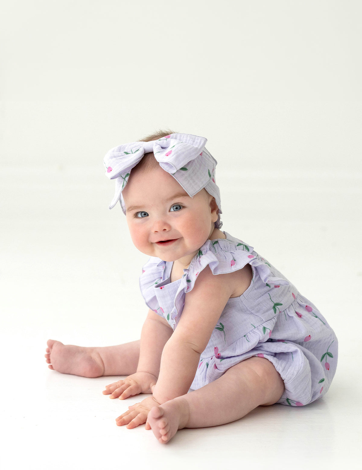 A smiling blue-eyed baby sits on the floor amid pink tulips, wearing the Mabel and Honey Tiny Tulip Baby Romper with ruffles and a matching cherry-print bow headband, set against a white background.